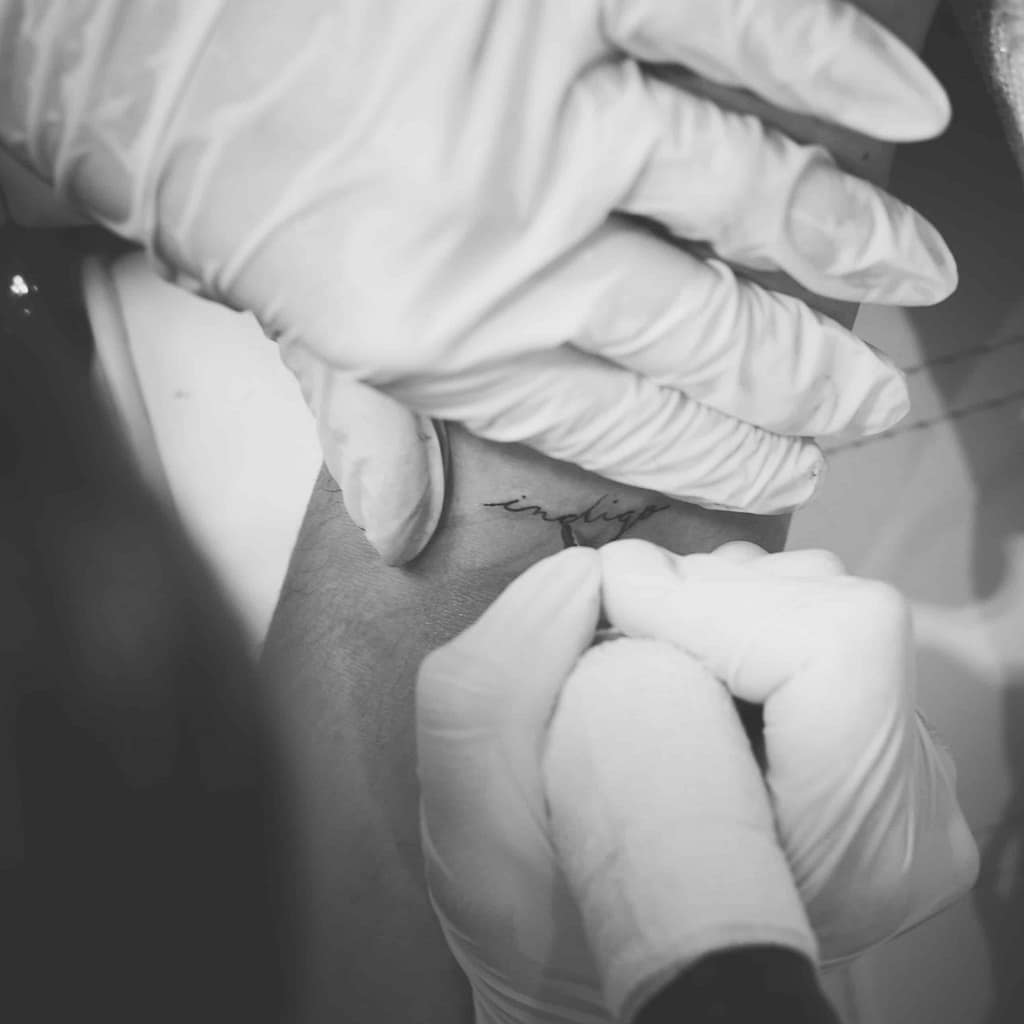 Tattoo artist inking the word 'Indigo' on inner arm as a symbol of spiritual identity and sensitivity — permanent mark of Indigo soul embodiment and self-acceptance.