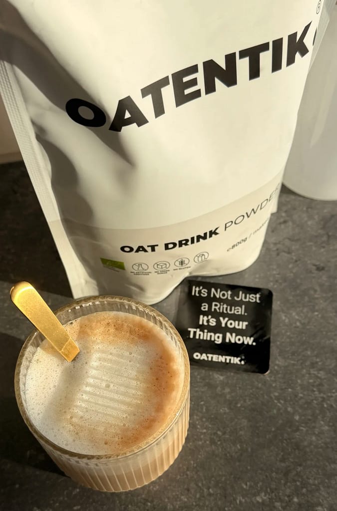 Frothy oat milk coffee in a textured glass, made with OATENTIK® oat drink powder, with branded packaging and a black inspirational card reading 'It's Not Just a Ritual. It's Your Thing Now' on a stone countertop in morning sunlight